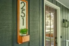 Modern house number 251 displayed on a vertical sign beside a front door, with a small potted plant underneath on a wooden shelf.