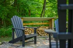 A peaceful outdoor scene with a black Adirondack chair in the foreground and a woodpile under a shelter in the background, surrounded by green trees.