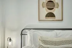 Modern bedroom with a decorative wall hanging above the bed, white linens, and a bedside lamp.
