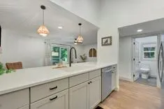 Modern kitchen interior with white cabinets, quartz countertops, and pendant lighting, connected to a small bathroom.