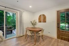 Bright dining room with a round wooden table, modern chairs, and open sliding doors leading to a deck with greenery.