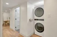 Modern laundry room with stacked washer and dryer next to a small wire-basket shelf, with an open door revealing a dining area in the background.