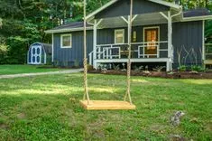 Wooden swing hanging from a tree in a lush backyard with a dark gray house and a small garden shed in the background.