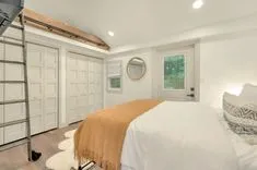 Bright modern bedroom with white walls, a large bed with a tan blanket, built-in closets, round mirror, and a loft ladder.