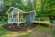 Cozy grey cabin with white trim, front porch, and adjoining wooden deck surrounded by green forest.