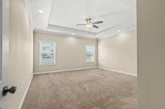 Empty room with beige carpeting, white walls, ceiling fan, and windows allowing natural light.