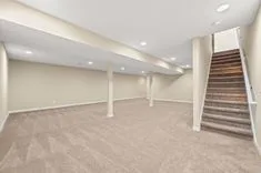 Empty basement interior with beige carpeting, white walls with several support columns, and a staircase to the upper level.