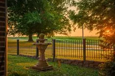 Sunset view through a wrought iron fence with a stone bird bath in the foreground and a green field in the background.