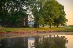 Brick house surrounded by trees with a reflective pond in the foreground during sunrise.