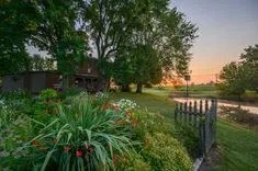 Sunrise over a serene rural landscape with a brick house, lush garden, green lawn, a pond, and a wooden fence.