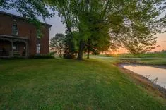 Sunrise view beside a brick house with a porch, large green tree, manicured lawn, and a small pond.