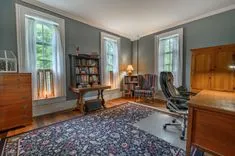 Traditional home office with a wooden desk, bookshelf, classic armchair, and oriental rug, bathed in natural light from tall windows.