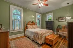 Elegant traditional bedroom with hardwood floors, a wooden bed with floral bedding, a black armoire, an armchair, and a green color scheme with natural light coming through the windows.