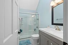 Modern bathroom interior with light blue walls, featuring a glass shower enclosure, white vanity with sink and bronze faucet, large mirror with dark frame, and white toilet.