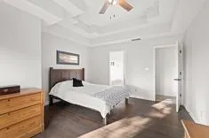 Bright and airy bedroom with a dark wood bed frame, white bedding, a unique tiered ceiling design with a ceiling fan, hardwood flooring, and an open door leading to another room.