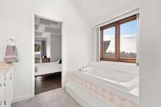 Bright bathroom interior with a built-in bathtub featuring decorative tile detailing, adjacent to a bedroom visible through an open door, and a window looking out to a rural landscape.