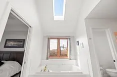 Bright bathroom interior with a built-in bathtub under a window offering a countryside view, a skylight, and adjacent to a bedroom visible through an open door.
