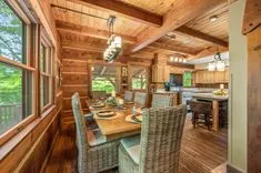 Cozy wooden cabin interior with a dining table set for a meal, rustic kitchen with modern appliances, and large windows showing greenery outside.
