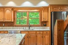 A bright kitchen interior with wooden cabinets, a granite countertop, and a window overlooking green foliage.
