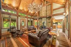 Spacious rustic living room with a vaulted wooden ceiling, stone fireplace, large chandelier, leather furniture, and hardwood floors leading to an open kitchen.