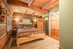 Cozy rustic bedroom with wooden walls, ceiling beams, and a bed with decorative animal-themed bedding, adjacent to a hallway with a wooden door.