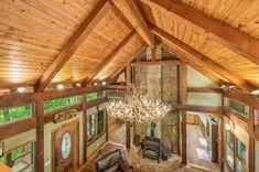 Interior view of a rustic cabin with a high wooden ceiling, exposed timber beams, antler chandelier, stone fireplace, and kitchen area in the background.