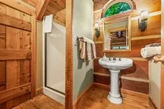 Interior of a rustic bathroom with wood paneling, featuring a shower stall, pedestal sink, and towels hanging on the wall.