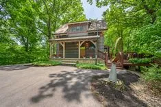 Charming two-story house with a front porch and deck surrounded by lush green trees, featuring a unique metal sculpture in the yard.