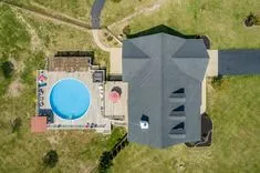 Aerial view of a house with a gray roof, adjacent circular blue swimming pool, and surrounding lawn area.