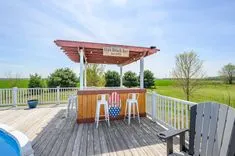 Outdoor wooden deck with a bar area, stools, and a pergola, overlooking a grassy field under a clear blue sky.