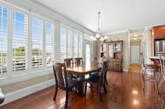 Bright dining room with a wooden table, chairs, and plantation shutters on windows with a view into a kitchen with bar stools.
