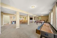 Spacious basement recreation room with carpeted flooring, a shuffleboard table, columns, and ample lighting with areas for dining and relaxation in the background.