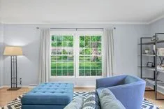 Bright living room interior with blue ottoman, sofa, patterned rug, floor lamp, shelving unit, and a large window with a view of green foliage.