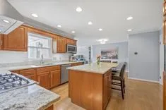 A modern kitchen with wooden cabinets, granite countertops, stainless steel appliances, hardwood floors, and a center island with bar stools.
