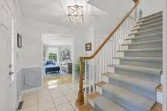 Interior view of a clean, bright entryway with tiled flooring and a wooden staircase leading up; living area visible through the doorway.