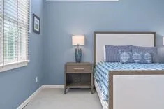 A neatly organized bedroom with light blue walls, a double bed with patterned bedding, a wooden nightstand with a lamp, and a window with blinds.