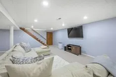 Modern basement living area with sectional sofa, flat-screen TV, and stairway.