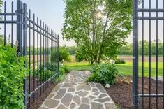 Open wrought iron gate leading to a stone pathway and lush garden with a large tree and green lawn in the background.