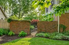 A lush garden with a brick wall and iron gate flanked by green shrubs, with a house partially visible to the left.