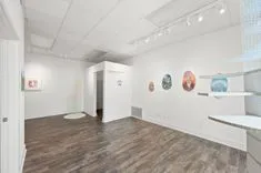 Interior of a bright art gallery with white walls displaying various artworks and a wooden floor.