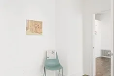 A minimalist room with white walls, a simple green chair against the wall, and a framed artwork hanging above it.