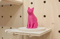Bright pink cat sculpture displayed on a white shelf against a pegboard wall.