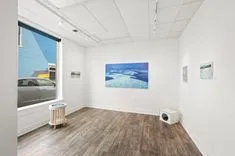 Interior of a small art gallery with paintings displayed on white walls and a projector on the floor.