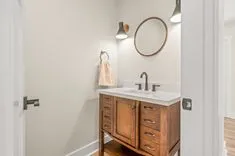 Modern bathroom interior with wooden vanity cabinet, white countertop, round mirror, and wall sconce.