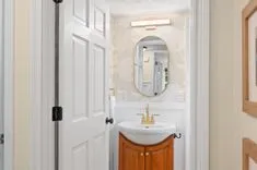  A view into a small bright bathroom with an oval mirror above a vanity with a wooden cabinet and a vessel sink, seen through an open door.