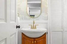 A small bathroom with white walls, featuring an elegant pedestal sink with a wooden cabinet and a gold faucet underneath an oval mirror.