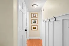 A narrow hallway with white walls, multiple doors, framed pictures on the wall, and a red floor mat.