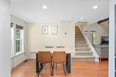 Bright and spacious dining room with a wooden table, two chairs, hardwood floors, and a staircase in the background.