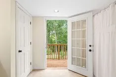 Bright interior room with open French doors leading to a wooden deck, surrounded by trees.