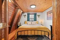 Cozy attic bedroom with a slanted wooden ceiling, queen-size bed with yellow bedding, wall-mounted plants, and a framed landscape painting.